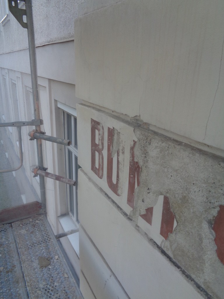 Fassade mit abgeplatztem Putz und freigelegtem Beton am Schriftzug vor der Sanierung
